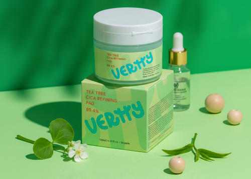 VERRTY Tea Tree Skin Refining Pad set displayed on a light green surface with a green backdrop. The set includes a white jar with a light green label, its matching light green box, a small dropper bottle with a gold collar, and decorative green leaves and peach-colored beads.