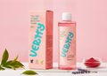 A bottle and box of Verity Tea Tree Vitamin Boosting Toner with pink liquid, styled on a white surface with green leaves, lavender, and a small bowl of red powder against a pink background.